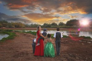 A family of five in formal outfits, including a mother in a red dress and a girl in green, stands by a pond at a colorful sunset.