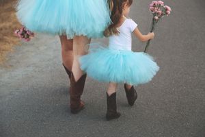 A mother and daughter in matching turquoise tutus and cowboy boots walk along a path holding pink flowers.