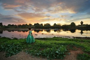 A young girl in a flowing green gown stands beside a calm reflective pond under a vivid orange and purple sunset sky.