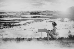 Black and white photo of a little girl in a tutu playing a small piano set in a misty, watery field.