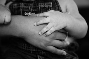 Black and white close-up of a baby's small hand resting on a parent's arm, with the child wearing plaid overalls.