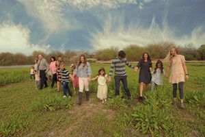 Family members of all ages hold hands and walk through a grassy field beneath a blue sky with wispy white clouds.