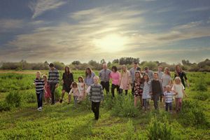 Extended family stands together in a green meadow at sunset, with glowing sunlight breaking through the clouds above.