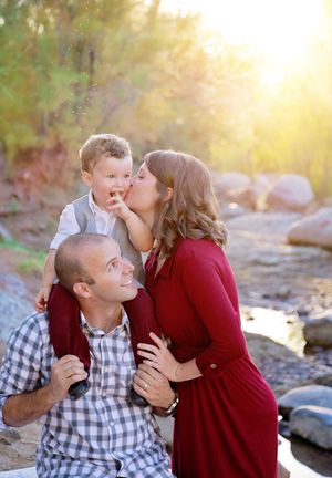 A smiling father carries a toddler on his shoulders while the mother kisses the child's cheek by a sunlit creek in autumn.