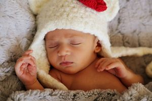 Newborn baby sleeping peacefully in a cream knit hat with a red accent, resting on a soft gray fur blanket.