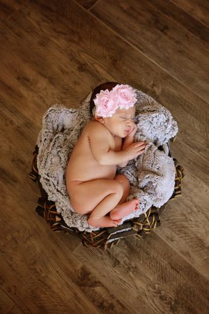 Overhead view of a newborn baby girl in a pink flower headband sleeping curled up in a basket on a rustic wood floor.