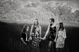 Black and white family portrait of a mother and three girls holding hands while walking through tall grass.