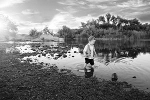 Black and white photo of a toddler wading barefoot through shallow lake water over rocks at sunset.