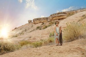 Young girl in a light dress stands on a sunlit desert hillside with rock formations and golden sunset glow.