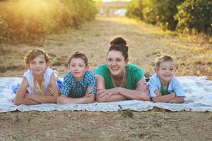 Mother and three smiling boys lie together on a blanket in a sunlit orchard during a warm family photo session.