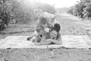 Black and white family photo of a laughing mother and three children piled playfully on a picnic blanket outdoors.