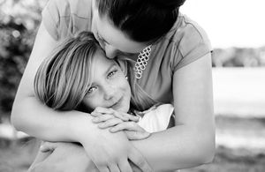 Black and white portrait of a mother hugging her smiling young daughter from behind in an outdoor setting.