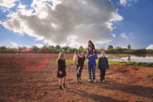 Family of five standing in an open field beside a pond under a blue sky with large billowing clouds.