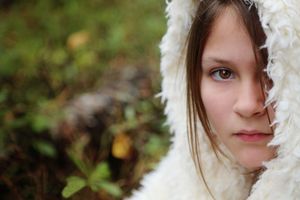 Close-up portrait of a young girl with long brown hair wearing a white fuzzy hood against a blurred green autumn background.