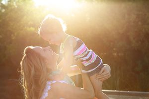 Mother lifting her smiling child high in warm golden sunlight with a glowing backlit outdoor background.