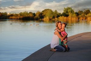 Mother kneeling on a lakeside path at sunset while hugging her young daughter, with ducks and golden trees in the background.