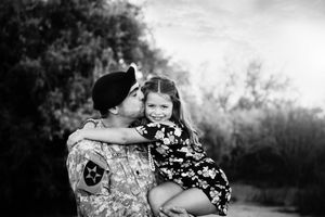 Black and white photo of a father in military uniform kissing his smiling daughter in a floral dress as she hugs him outdoors.