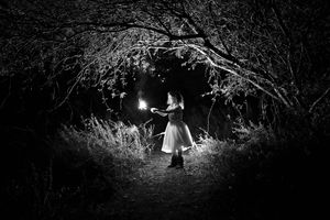 Black and white night photo of a girl in a white dress holding a sparkler on a path beneath an arch of tree branches.