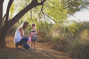 Mother in jeans kneels beside a little girl in a striped dress under a tree on a sunlit trail with tall grass.