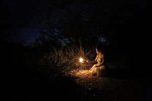 Girl in a light dress sitting in the dark outdoors, softly lit by a glowing sparkler among bushes and trees at night.