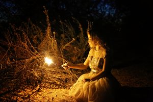 Girl in a lace dress holding a sparkler beside dry brush at night, with warm golden light glowing against the dark background.