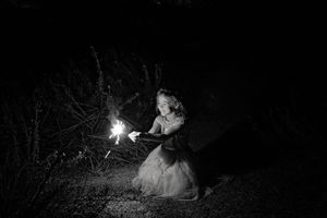 Black and white night portrait of a smiling girl in a tulle dress crouching and holding a bright sparkler in a dark field.