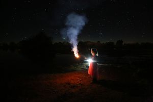 Girl in a red dress holding a sparkler under a starry night sky as a plume of smoke rises beside her in a dark field.
