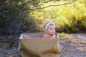 Smiling baby girl with a white headband sitting in a burlap bin outdoors among desert shrubs and green foliage.