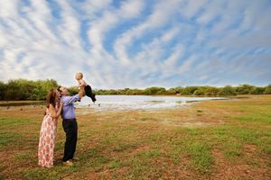 Parents lifting a laughing toddler into the air in a grassy field beside a lake under a dramatic blue sky with wispy clouds.