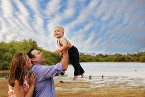 Smiling father lifts a laughing toddler into the air beside a mother, with a lake and dramatic cloudy sky behind them.