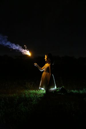 Girl in a sparkly dress holds a lit firework in a dark grassy field at night, with trailing smoke and glowing light.