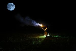 Girl in a dress kneels in a grassy field holding a smoking sparkler under a bright full moon in the dark night sky.