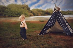 Toddler in dark suspenders walks across a grassy lakeside field beside a polka dot teepee under a cloudy blue sky.