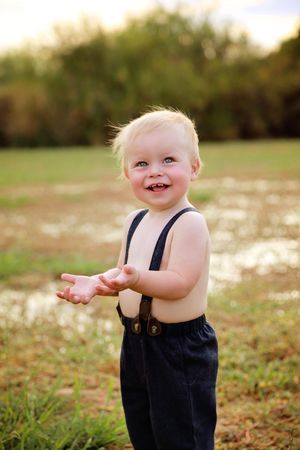 A smiling blond toddler boy in navy suspenders and trousers claps his hands in a grassy field with soft golden light.