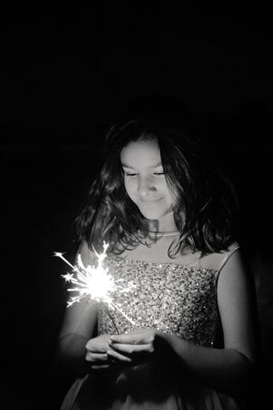 Black and white portrait of a smiling girl in a sequin dress gazing down at a bright sparkler held in her hands.