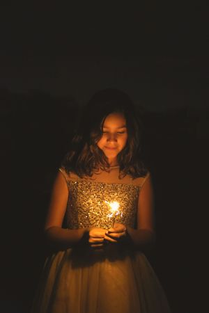Girl in a gold sequin dress smiles down at a small glowing sparkler held in her hands against a dark background.