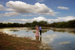 Couple stands close together at the edge of a reflective lake under a big blue sky with fluffy white clouds.