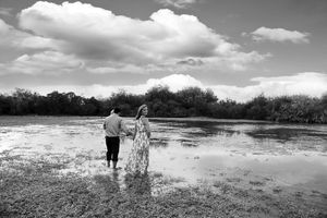 Black and white photo of a couple holding hands as they walk through shallow lake water under a cloudy sky.