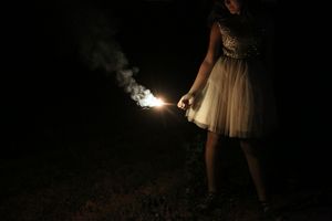 Girl in a tulle skirt and sequin top holds a lit sparkler that glows and smokes in the darkness at night.
