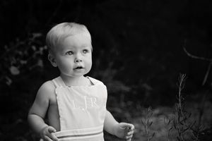 Black and white portrait of a blond toddler in a monogrammed romper standing outdoors and looking up with curiosity.