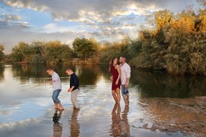 Two boys and a couple wade barefoot in shallow lake water at sunset, with golden trees and a colorful cloudy sky behind them.