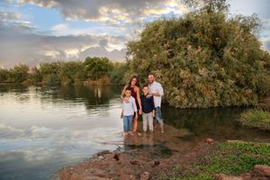 A smiling family of four poses at the edge of a calm lake at dusk, surrounded by lush green trees and a pastel sky.