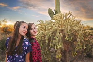Two smiling teenage girls pose close together beside a cholla cactus and tall saguaro against a warm desert sunset sky.