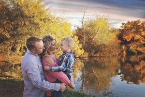Parents embrace their young son by a calm lake reflecting colorful autumn trees in a warm family portrait at sunset.