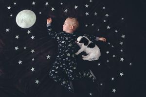 A sleeping toddler in star-print pajamas lies beside a tiny puppy on a dark backdrop decorated with glowing stars and a moon.