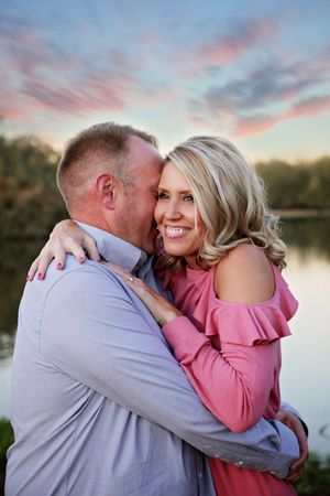 A smiling couple embraces by a lake at sunset, the man nuzzling the blonde woman in a pink ruffled top against a pastel sky.