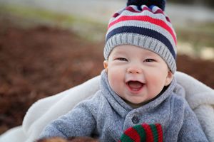 A laughing baby in a striped knit beanie and gray jacket sits on a soft white blanket outdoors on an autumn day.