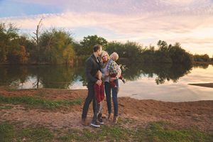 A family of four shares a loving embrace on a lakeshore at sunset, with trees reflected in the calm water behind them.