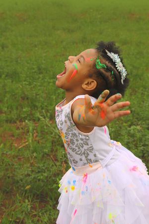 A joyful girl with paint-smeared face and arm outstretched cries out in a grassy field, wearing a white tutu skirt and flower headband.