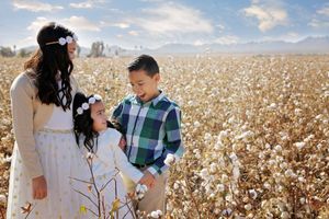 A woman in a cream cardigan and two children in plaid and white pose together in a cotton field under a blue sky.